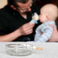 Vater spielt mit Baby am Tisch, während dieses passiv den Rauch einer auf dem Tisch im Aschenbecher glühenden Zigarette inhaliert.